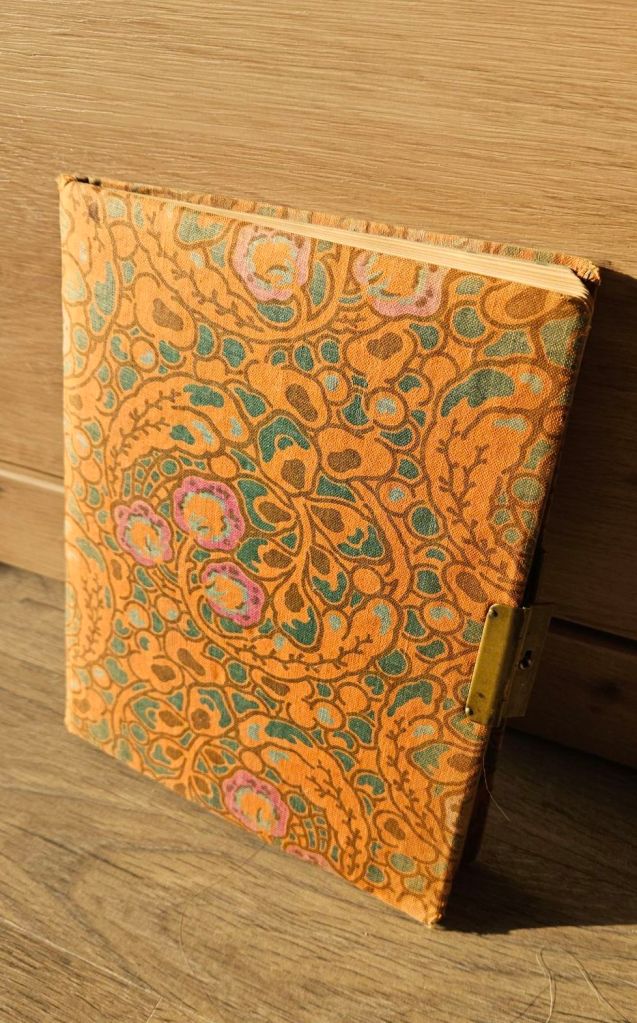 Ein etwa A5 großes orangenes Buch, mit braunem Blumenmuster. Man kann erkennen, dass es alt und ziemlich abgegriffen ist.