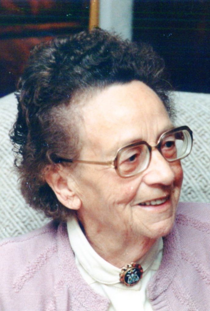 Ein älteres Bild aus den 80er Jahren. Es zeigt eine alte Frau von etwa 80 Jahren. Sie hat kurze dunkle Haare, eine große Brille, eine weiße Bluse mit einer Brosche am Kragen und einer rosa Strickjacke drüber. Sie lacht fröhlich zur rechten Seite.