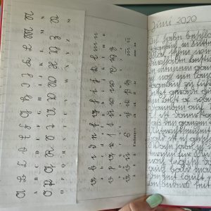 Ein Ausschnitt von einem aufgeschlagenem Buch. Die rechte Siete zeigt das Sütterlin Alphabet und links sieht man abgeschnitten einen Tagebucheintrag vom Juni 2020 in großen Sütterlin Buchstaben.